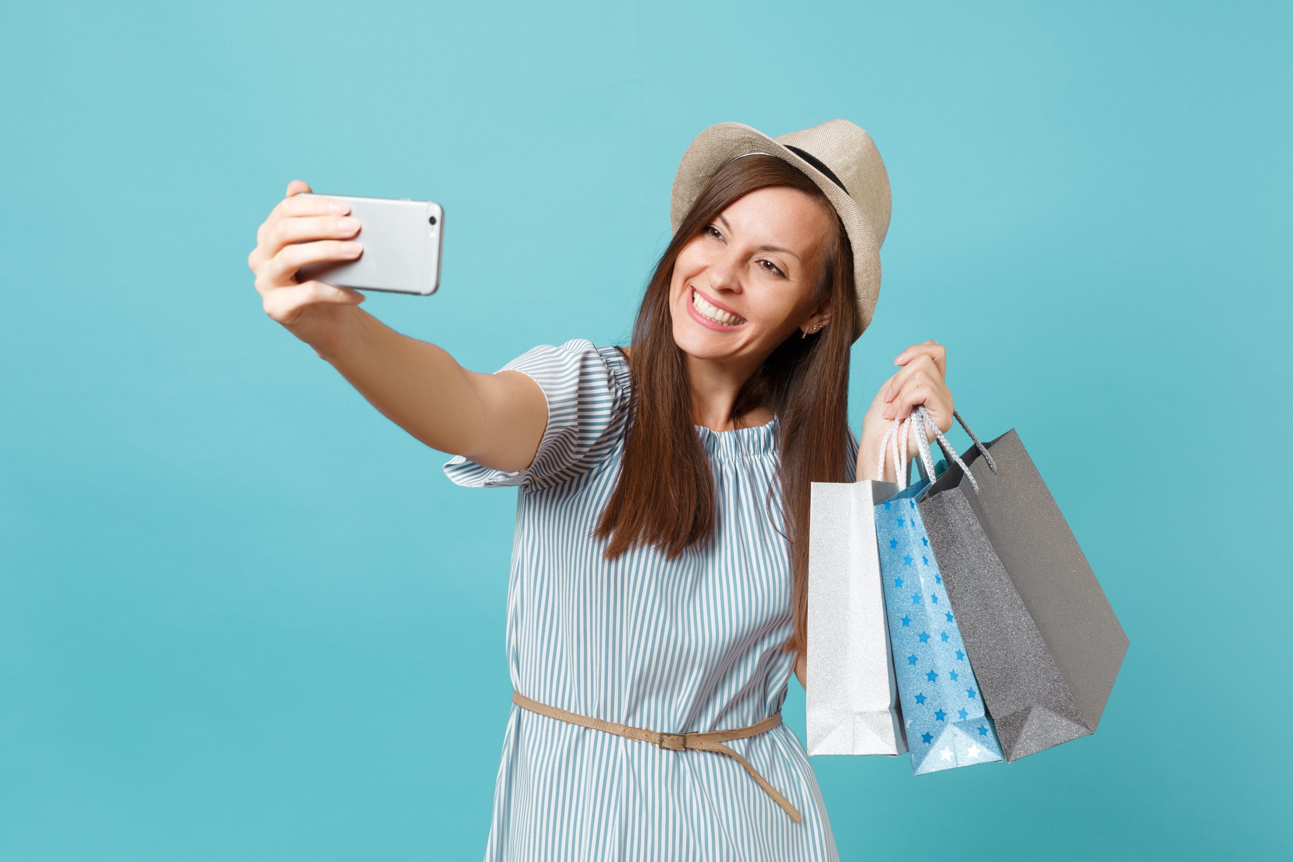 Portrait smiling woman in summer dress, straw hat holding packages bags with purchases after shopping doing selfie shot on mobile phone isolated on blue pastel background. Copy space for advertisement